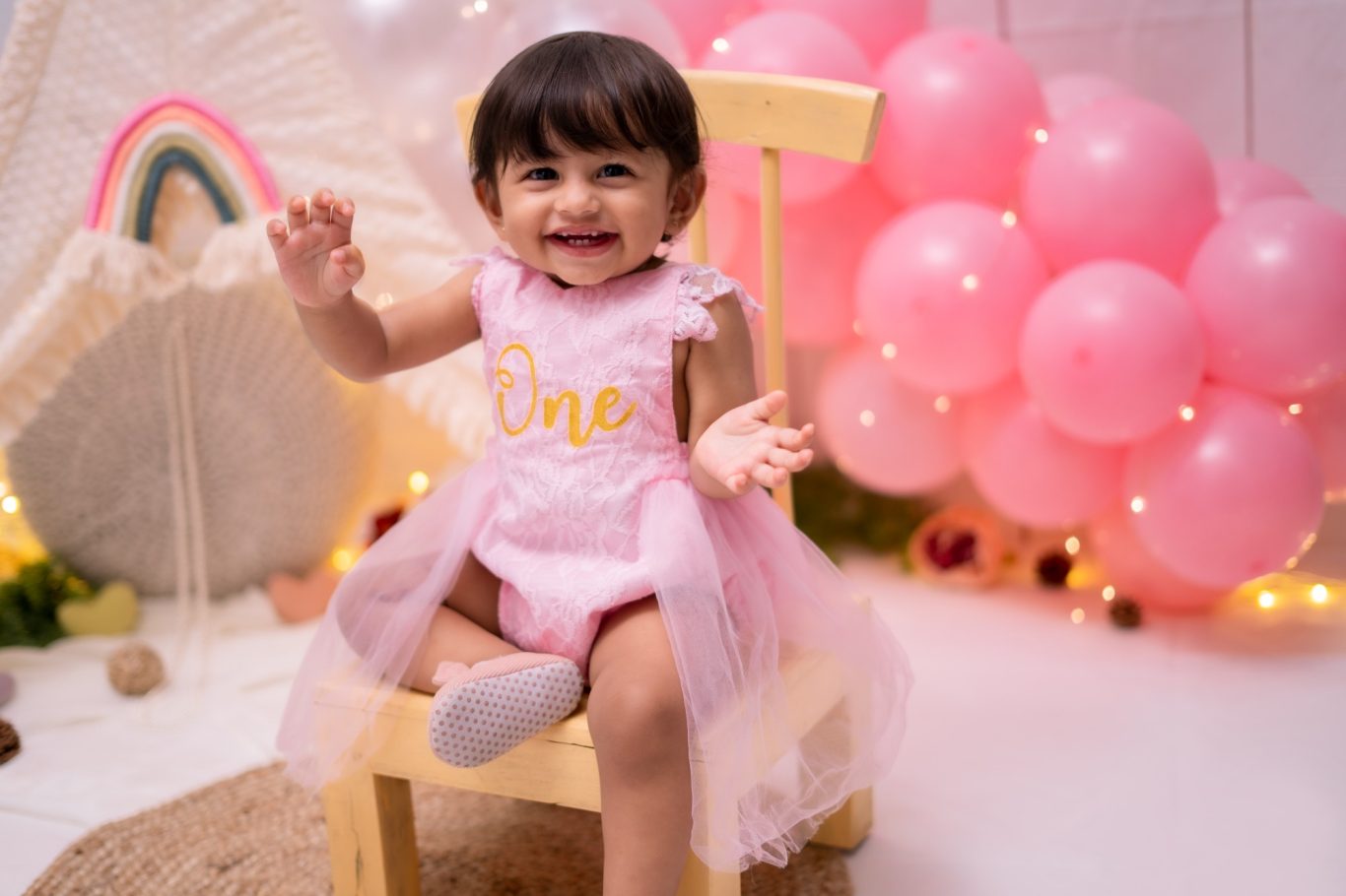 Smiling toddler in a pink dress sits on a chair surrounded by pink balloons.