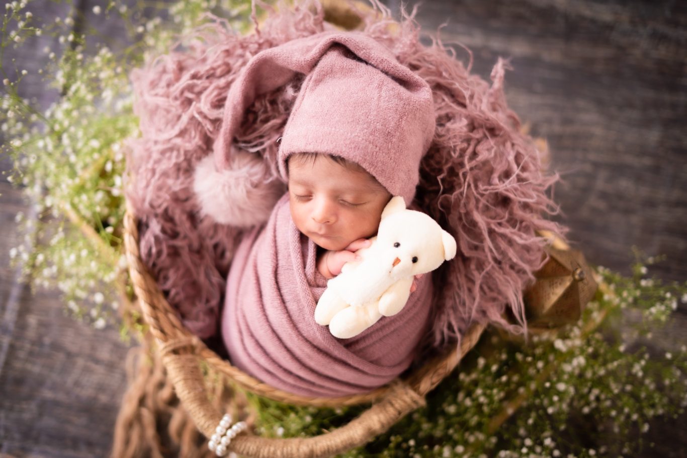 A swaddled baby in a pink hat, holding a teddy bear, nestled in a basket of greenery.