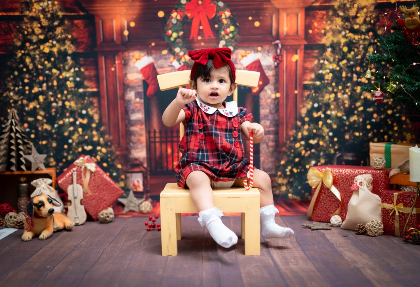 A baby in a red plaid outfit sits on a wooden chair surrounded by festive decorations.