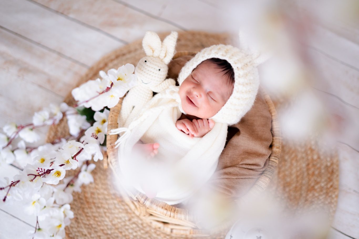 Smiling baby in a cream knitted outfit, with a soft toy and flowers around.
